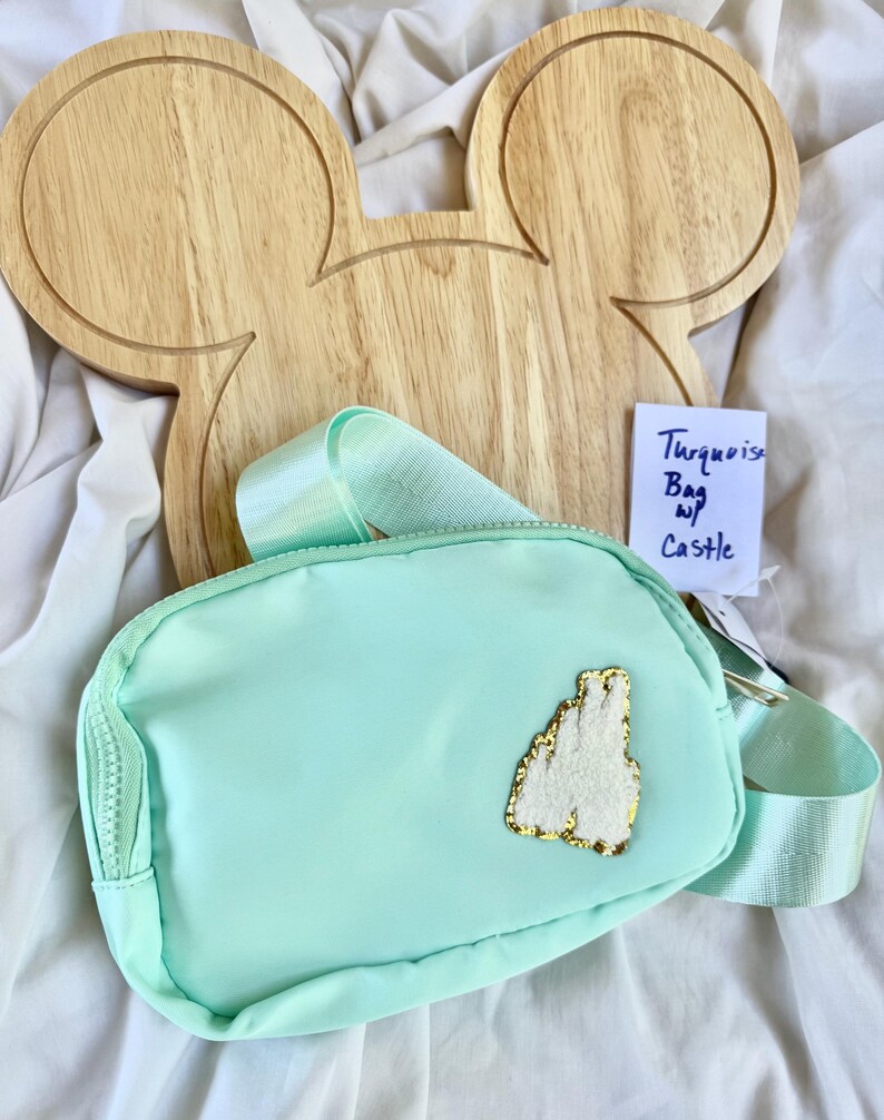 May include: A turquoise belt bag featuring a white textured castle patch with gold trim on the front. The bag has a matching turquoise adjustable strap and a zipper closure. A small white tag attached to the bag displays the handwritten text Turquoise Bag w/ Castle.