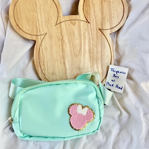 May include: A turquoise belt bag featuring a textured pink and white character head patch with a gold glitter border. The bag has a matching turquoise adjustable strap and a zipper closure. A small white tag reads Turquoise Bag w/ Pink Head.