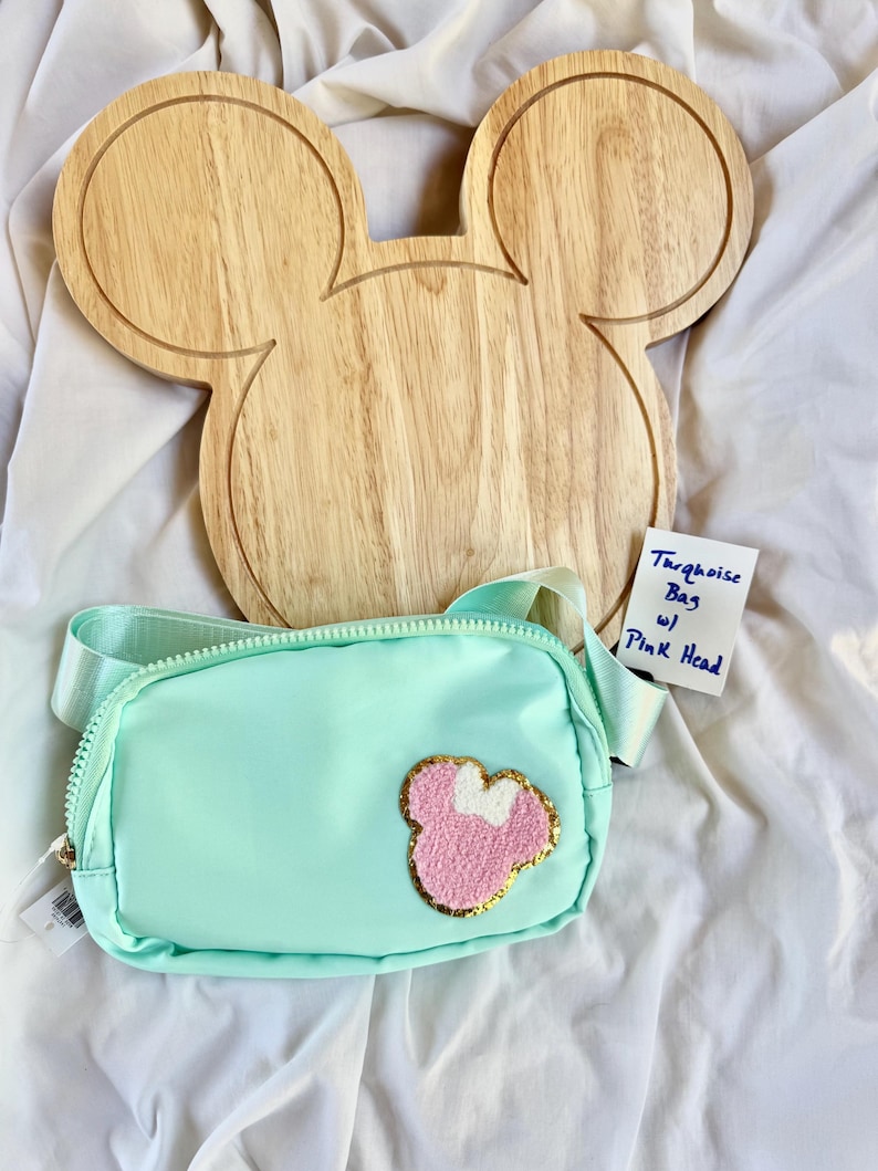 May include: A turquoise belt bag featuring a textured pink and white character head patch with a gold glitter border. The bag has a matching turquoise adjustable strap and a zipper closure. A small white tag reads Turquoise Bag w/ Pink Head.