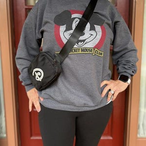 Puede incluir: Bolso bandolera negro con dise&ntilde;o de pretzel blanco. El bolso tiene una correa negra y cierre de cremallera. La persona lleva una sudadera gris con un gr&aacute;fico de Mickey Mouse Club y leggings negros.
