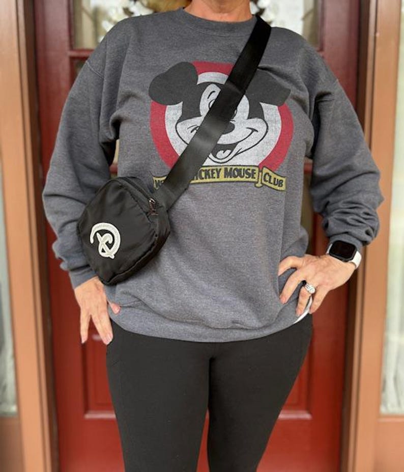 Puede incluir: Bolso bandolera negro con dise&ntilde;o de pretzel blanco. El bolso tiene una correa negra y cierre de cremallera. La persona lleva una sudadera gris con un gr&aacute;fico de Mickey Mouse Club y leggings negros.