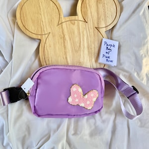 May include: A purple fanny pack featuring a textured pink bow with white polka dots on the front. The bag has a matching purple adjustable strap with a black plastic buckle. A small white note reads Purple Bag w/ Pink BOW.