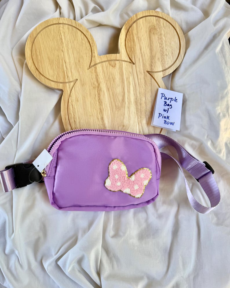May include: A purple fanny pack featuring a textured pink bow with white polka dots on the front. The bag has a matching purple adjustable strap with a black plastic buckle. A small white note reads Purple Bag w/ Pink BOW.