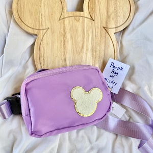 May include: A purple belt bag featuring a central cream-colored mouse head patch with a gold glitter border. The bag has a matching purple adjustable strap with a black plastic buckle. A small white tag displays the handwritten text Purple Bag w/ Mickey Head.