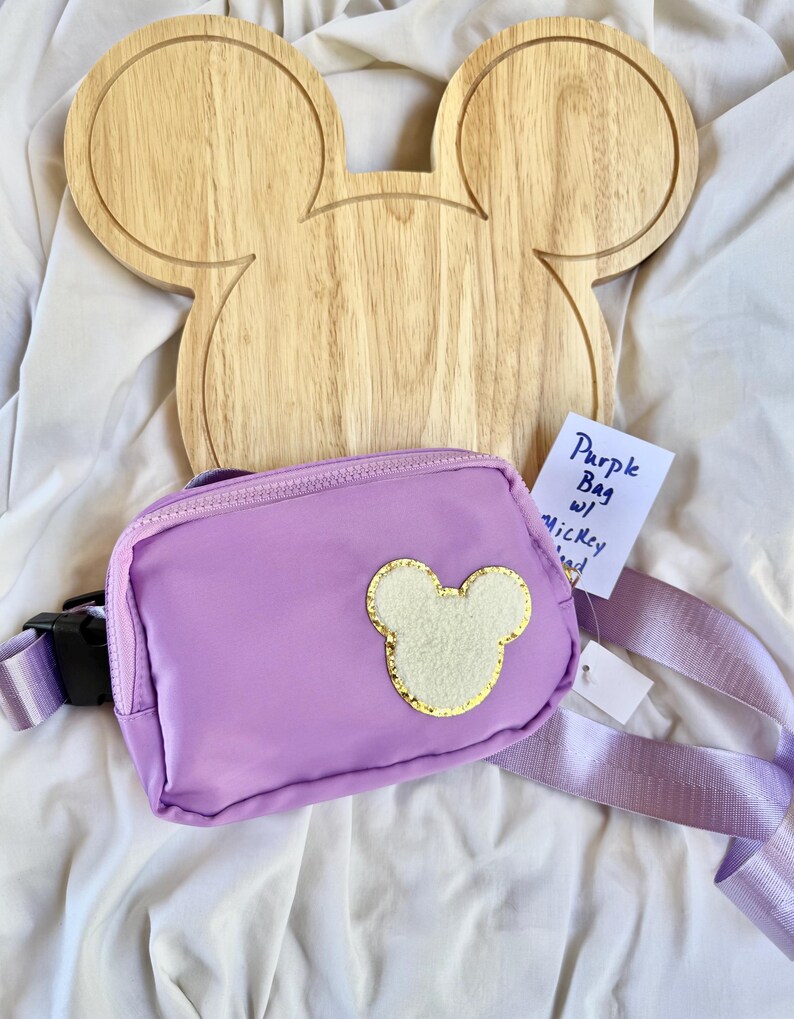 May include: A purple belt bag featuring a central cream-colored mouse head patch with a gold glitter border. The bag has a matching purple adjustable strap with a black plastic buckle. A small white tag displays the handwritten text Purple Bag w/ Mickey Head.
