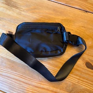 Puede incluir: Ri&ntilde;onera negra de nailon con un compartimento principal con cremallera y correa ajustable. La correa tiene una hebilla de pl&aacute;stico negro. La bolsa est&aacute; sobre una superficie de madera.