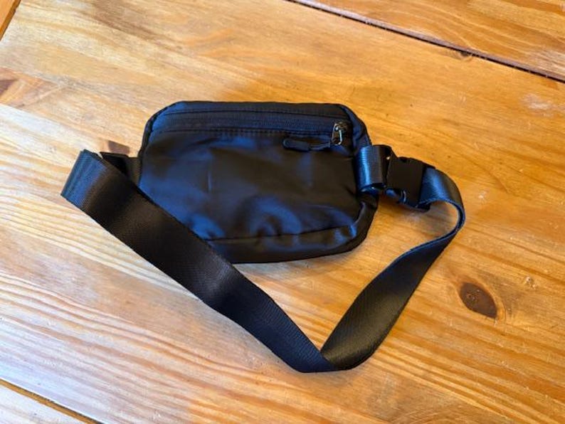 Puede incluir: Ri&ntilde;onera negra de nailon con un compartimento principal con cremallera y correa ajustable. La correa tiene una hebilla de pl&aacute;stico negro. La bolsa est&aacute; sobre una superficie de madera.