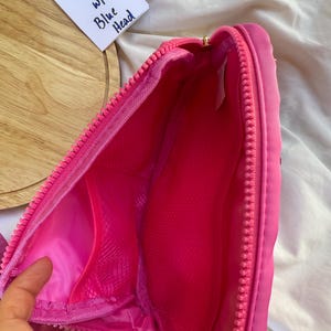 May include: A bright pink zippered bag featuring a main compartment with a mesh interior pocket. A white tag is visible with the handwritten text Pink bag w/ Blue Head. The bag has a thick pink zipper closure and a smooth, solid pink fabric exterior.