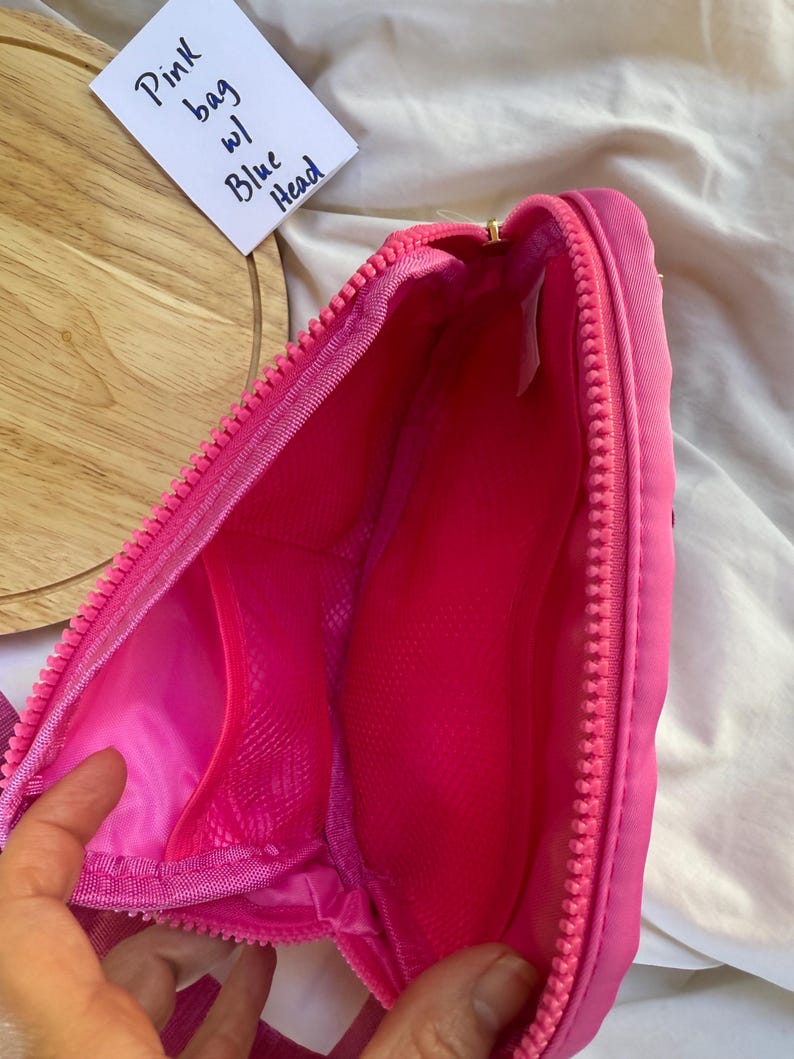 May include: A bright pink zippered bag featuring a main compartment with a mesh interior pocket. A white tag is visible with the handwritten text Pink bag w/ Blue Head. The bag has a thick pink zipper closure and a smooth, solid pink fabric exterior.