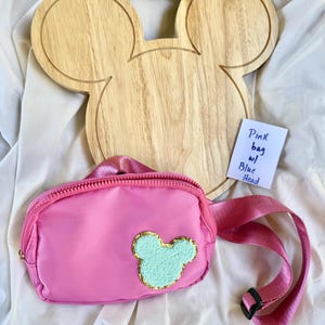 May include: A pink fanny pack with a light blue, textured, mouse-shaped patch outlined in gold glitter. The bag features a matching pink adjustable strap with a black plastic buckle. A small white note reads: Pink bag w/ Blue Head.