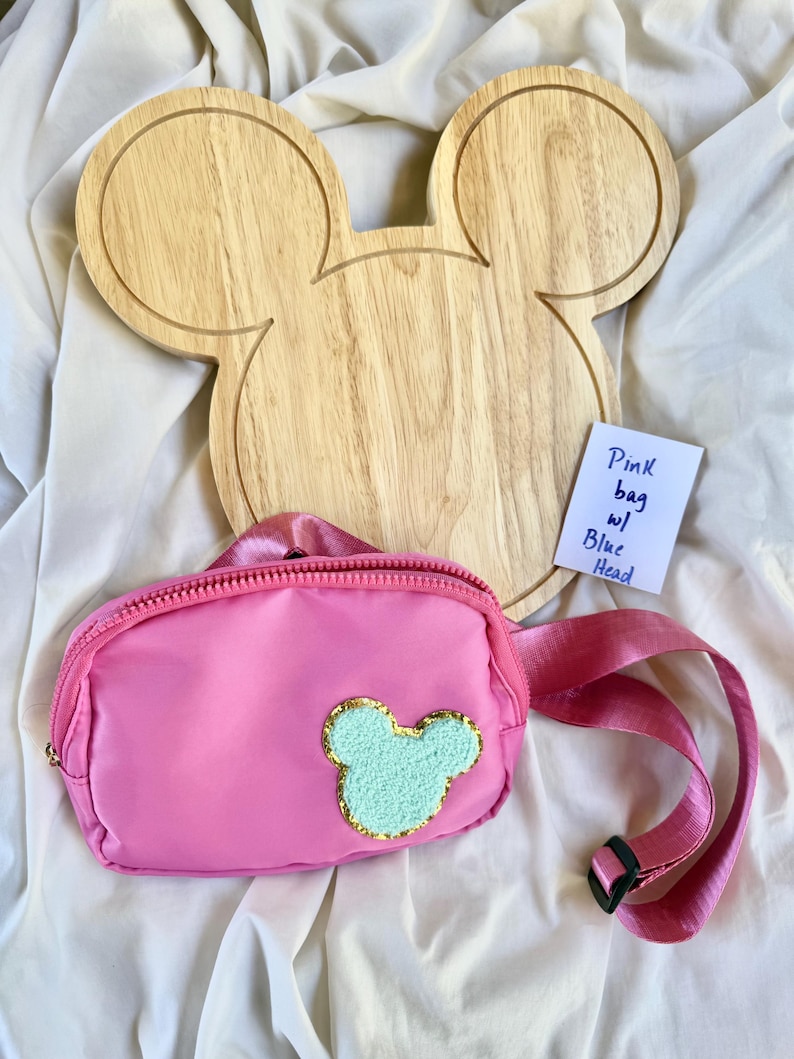May include: A pink fanny pack with a light blue, textured, mouse-shaped patch outlined in gold glitter. The bag features a matching pink adjustable strap with a black plastic buckle. A small white note reads: Pink bag w/ Blue Head.