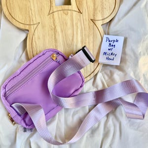 May include: A purple belt bag featuring a zippered main compartment and an adjustable strap with a black plastic buckle. A small white note is included with the handwritten text Purple Bag w/ Mickey Head, placed near a wooden Mickey head cutout.