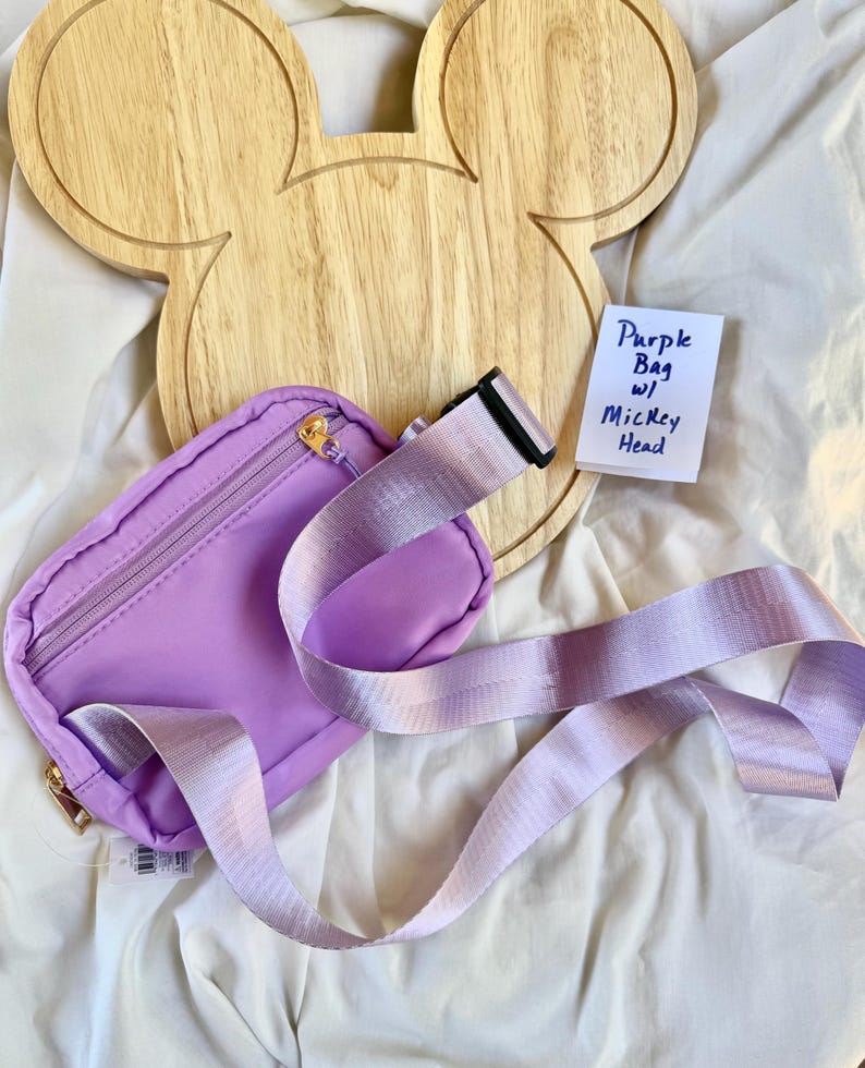 May include: A purple belt bag featuring a zippered main compartment and an adjustable strap with a black plastic buckle. A small white note is included with the handwritten text Purple Bag w/ Mickey Head, placed near a wooden Mickey head cutout.
