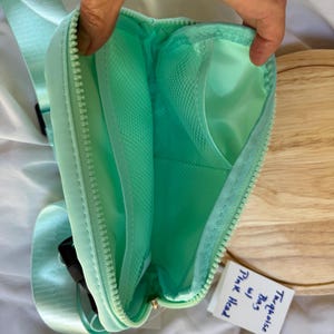 May include: A mint green crossbody bag with a zippered opening, featuring an interior mesh pocket and a matching adjustable strap. A small white tag is attached to the bag with the handwritten text "Turquoise Bag w/ Pink Head".