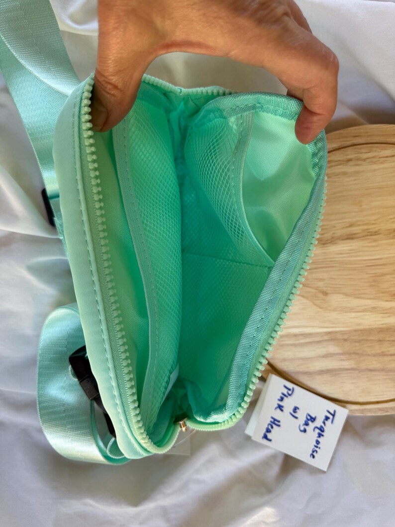 May include: A mint green crossbody bag with a zippered opening, featuring an interior mesh pocket and a matching adjustable strap. A small white tag is attached to the bag with the handwritten text "Turquoise Bag w/ Pink Head".