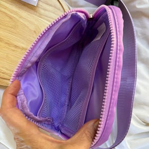 May include: A lavender belt bag with a matching zipper and an adjustable strap. The interior features a smooth lining and two mesh pockets. A small white tag is visible inside, and a paper note reads Bag w/ Pink Bow.