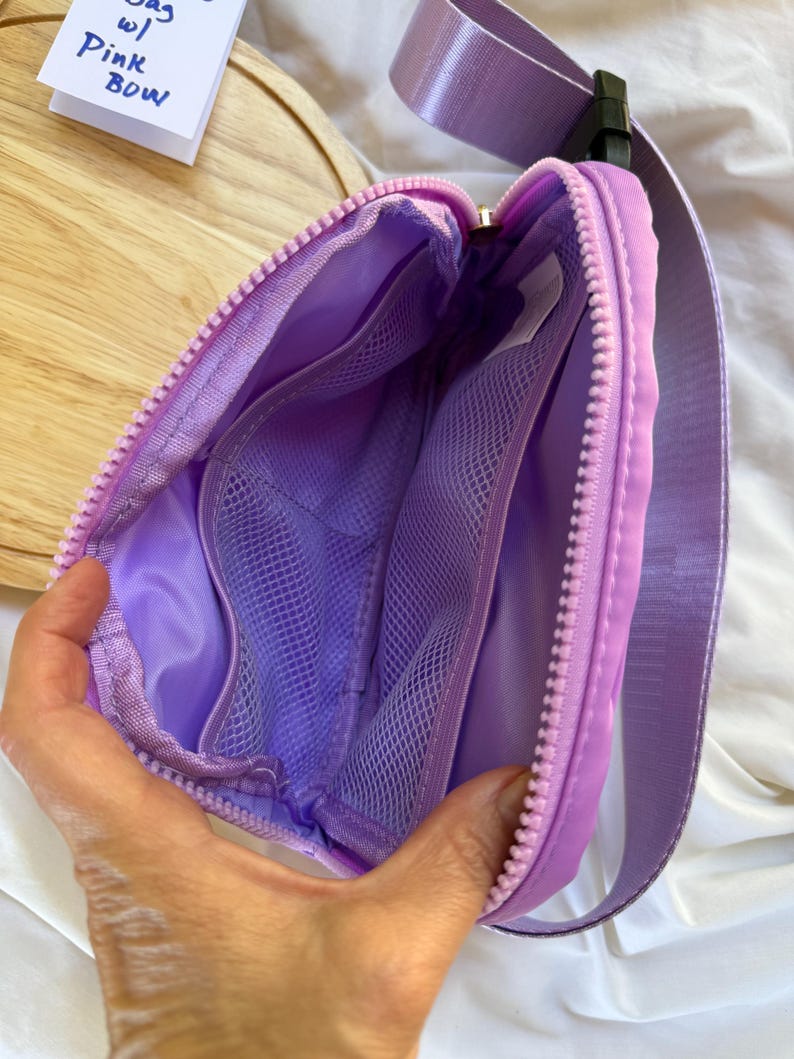 May include: A lavender belt bag with a matching zipper and an adjustable strap. The interior features a smooth lining and two mesh pockets. A small white tag is visible inside, and a paper note reads Bag w/ Pink Bow.