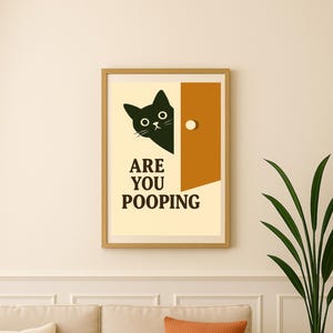 Peut inclure: Impression encadrée avec un chat noir regardant par une porte, avec le texte "ARE YOU POOPING". L'œuvre est dans un cadre en bois, au-dessus d'un canapé beige avec des coussins orange et crème.
