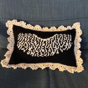May include: A rectangular black crochet pillow with a white geometric design in the center. The pillow has a scalloped, off-white border. The pillow is resting on a blue surface.