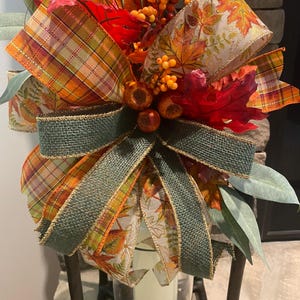 May include: A decorative autumn-themed bow featuring layers of plaid, burlap, and leaf-patterned ribbons in shades of orange, red, green, and gold. The arrangement is accented with artificial orange berries, red maple leaves, and green eucalyptus foliage.