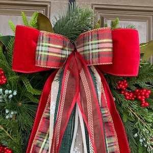 May include: A decorative wreath featuring lush green pine branches, clusters of bright red berries, and a large multi-layered bow. The bow combines solid red velvet ribbon, red and green plaid fabric, and long hanging streamers with snowflake patterns.
