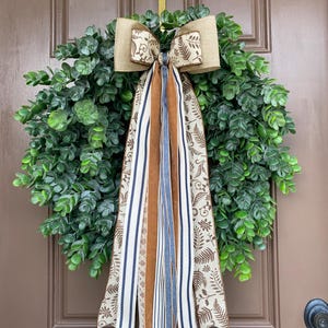 May include: A round wreath made of green eucalyptus leaves featuring a large decorative bow at the top. The bow and long, hanging ribbons are made of tan, cream, and navy blue fabric with patterns of brown leaves and vertical stripes.