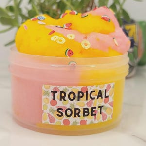 May include: A clear plastic container filled with yellow and pink slime. The slime is topped with a yellow and pink swirl, decorated with small fruit-shaped sprinkles. The container has a label that reads "TROPICAL SORBET".
