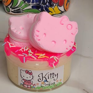 Pode incluir: Um frasco de vidro transparente cheio de uma substância amarela semelhante a creme, coberto com glacê rosa e granulado colorido. Dois biscoitos rosa em forma de Hello Kitty estão em cima do glacê. O frasco tem um rótulo que diz "Kitty Cookies".