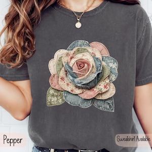 May include: A charcoal gray short-sleeve t-shirt featuring a large, central graphic of a multi-layered rose. The rose is composed of various fabric-like textures in shades of blue, pink, green, and cream, with visible stitching along the petal edges.