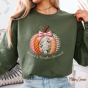 May include: A military green sweatshirt featuring a central graphic of a pumpkin with segments in orange polka dot, floral, and gingham patterns. A pink bow sits atop the pumpkin, which is printed with the text "blessed & Pumpkin Obsessed".