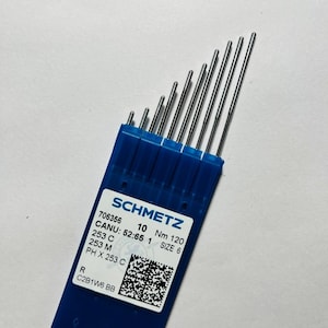 May include: A set of silver-colored sewing machine needles arranged in a blue plastic case. The label reads SCHMETZ, 706356, 10, Nm 120, SIZE 6, CANU: 52:65 1, 253 C, 253 M, PH X 253 C, R, C2B1W6 BB, and includes a square QR code graphic.
