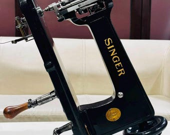May include: A black Singer sewing machine arm with gold lettering and a metallic top mechanism. It features a wooden handle, a circular gold emblem, and a black wheel at the base. The machine is positioned at an angle, showcasing the intricate metal components and the brand name printed vertically in gold on the side.