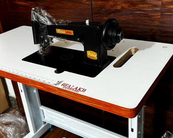 May include: A black industrial sewing machine with Falak Crown branding mounted on a white tabletop with a wood-grain edge. The machine features a blue foot pedal labeled Hi-Tech and a white metal stand. The Falak Enterprises logo is printed on the table.