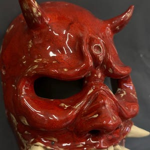 May include: A glossy red ceramic mask with two horns, large eye holes, and a wide mouth revealing white teeth. The mask has a textured surface with a slightly distressed appearance, suggesting an aged or handcrafted quality. The mask is likely for decorative or ceremonial purposes.