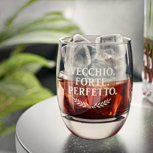 May include: A clear glass tumbler filled with ice cubes and a dark amber liquid. The glass has the words "VECCHIO. FORTE. PERFETTO." etched on the front, along with a decorative sprig design. The glass sits on a white surface.