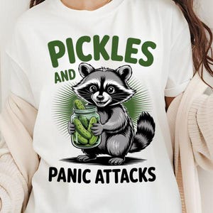 May include: White t-shirt featuring a cartoon raccoon holding a jar of pickles. The text above the raccoon reads "Pickles" and "Panic Attacks" is below. The raccoon is grey and black with a green background.
