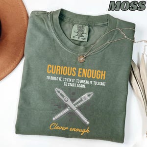 May include: A moss green t-shirt with the text "CURIOUS ENOUGH" and "Clever enough" in yellow. The shirt features a graphic of crossed pliers and circuit board elements. A silver necklace with a pendant is draped across the shirt.