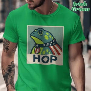 Puede incluir: Camiseta verde con un diseño de rana patriótica. La rana lleva un collar azul con estrellas blancas y una bufanda a rayas rojas y blancas. La palabra "HOP" está impresa en letras blancas en la parte inferior.