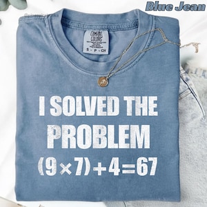 Puede incluir: Camiseta azul vaquero con el texto blanco "I SOLVED THE PROBLEM (9x7)+4=67". La camiseta tiene cuello redondo y mangas cortas. Un collar plateado con un colgante está sobre la camiseta.
