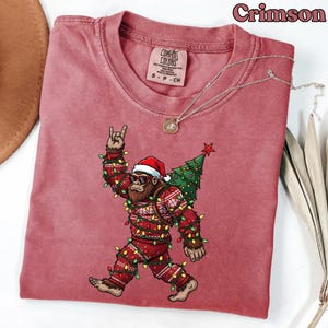 May include: Crimson-colored t-shirt featuring a graphic of a Bigfoot wearing a Santa hat, sunglasses, and a Christmas sweater, with a Christmas tree on its back. The Bigfoot is wrapped in Christmas lights, with the rock and roll hand gesture.