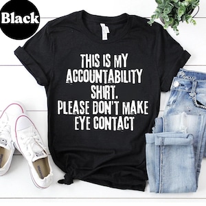 Puede incluir: Camiseta negra con el texto blanco "THIS IS MY ACCOUNTABILITY SHIRT. PLEASE DON'T MAKE EYE CONTACT". La camiseta se combina con vaqueros azules y zapatillas blancas. La palabra "Black" está en un círculo negro en la esquina superior izquierda.