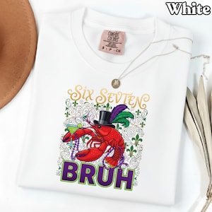 May include: White t-shirt featuring a red lobster in a top hat, holding a cocktail. The shirt displays the text "Six Sevten" in gold and "BRUH" in purple. A silver necklace and a Comfort Colors tag are also visible.