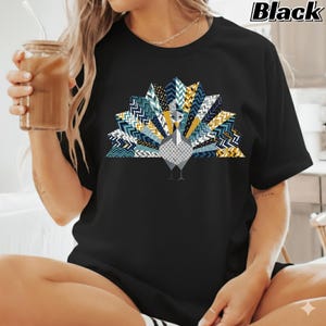 May include: Black t-shirt with a geometric peacock design. The peacock's tail feathers are a mix of blue, gold, and white chevron patterns. The shirt has the word "Black" printed in the upper right corner.