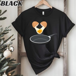 Puede incluir: Camiseta negra con un gráfico de un huevo roto y una sartén. La palabra "BLACK" está impresa en la parte superior. La camiseta está colgada de una percha de madera. El diseño tiene un tema culinario.