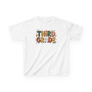 May include: White t-shirt with the words "THIRD GRADE" in a colorful, playful font. The text is in a mix of green, orange, yellow, and red, with decorative elements like lightning bolts and polka dots. A fun design for back-to-school.