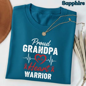 May include: Sapphire blue t-shirt with the text "Proud Grandpa of a Heart Warrior" in white and red. A gold necklace with a pendant is draped on the shirt. The design includes a heartbeat line and a red heart.