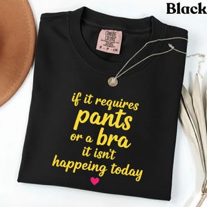 May include: Black t-shirt with yellow text reading "if it requires pants or a bra it isn't happening today". A small pink heart is below the text. A silver necklace with a pendant is on the shirt.