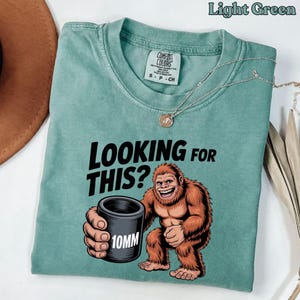 以下が含まれることがあります： 「LOOKING FOR THIS?」の文字と、黒い筒「10MM」を持った笑顔のビッグフットのイラストが描かれた、ライトグリーンのTシャツ。シルバーのネックレスがシャツの上にかけられています。