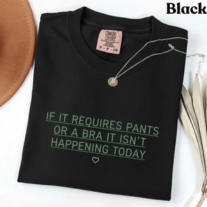 May include: Black crew neck t-shirt with the text "IF IT REQUIRES PANTS OR A BRA IT ISN'T HAPPENING TODAY" in green. A silver necklace with a pendant and a pink tag are visible. The shirt is a Comfort Colors brand.