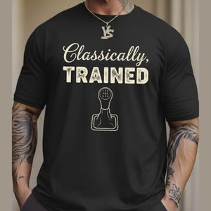 Może przedstawiać: Czarna koszulka z napisem "Classically, TRAINED" w stylu vintage, z grafiką dźwigni zmiany biegów samochodu. Koszulka ma okrągły dekolt.